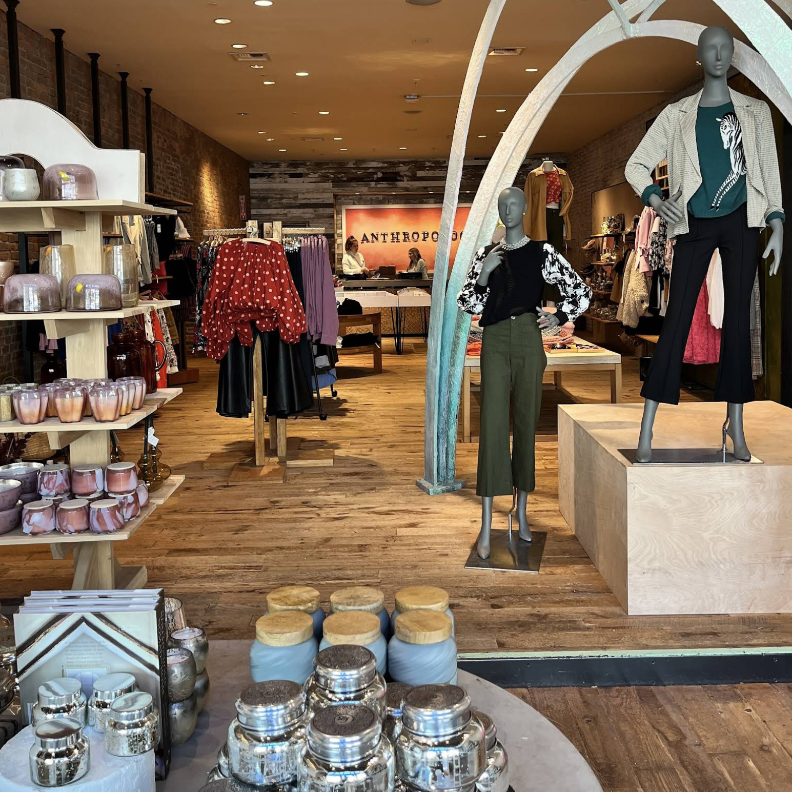 Anthropologie Ila Bridal Interior of a clothing boutique with mannequins in the foreground, racks of dresses and shelves, and a wooden floor; the store’s back wall reads ANTHROPOLOGIE.