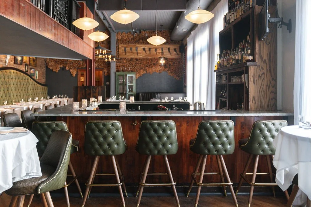 Cork Cow Ila Bridal Bar area with green tufted stools along a dark wooden counter in a warmly lit dining room.