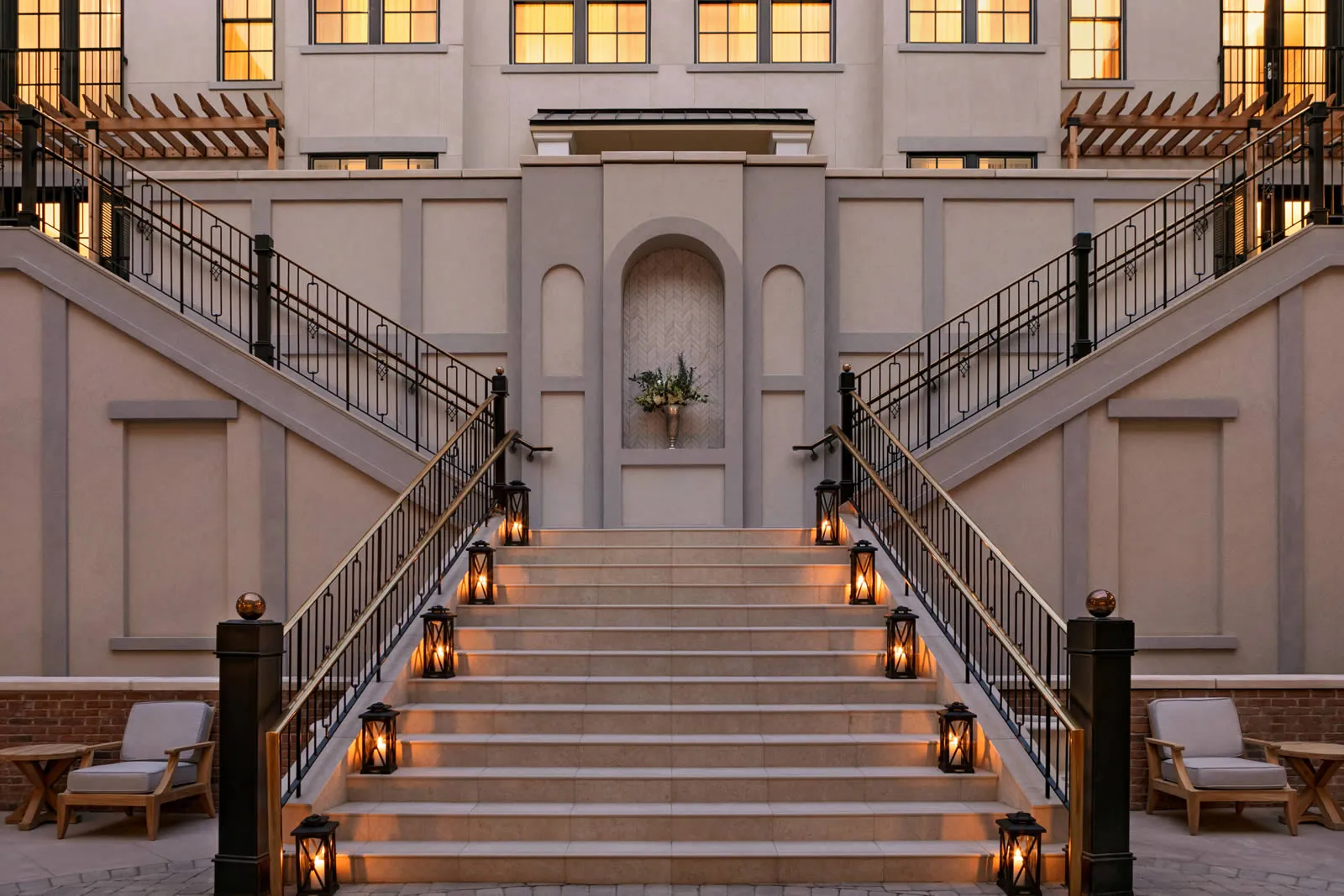 Symmetrical stone staircase leading to a central arched niche with a vase of greenery, flanked by black iron railings and lanterns along the steps.