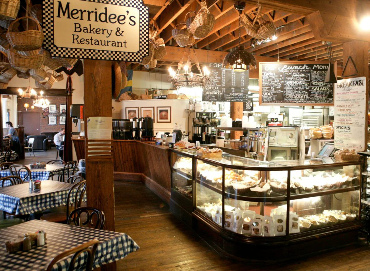 Merridees Bakery Restaurant Cropped Cozy bakery cafe interior with a glass pastry display, wooden beams, and blue-and-white checkered tables at Merridee's Bakery & Restaurant