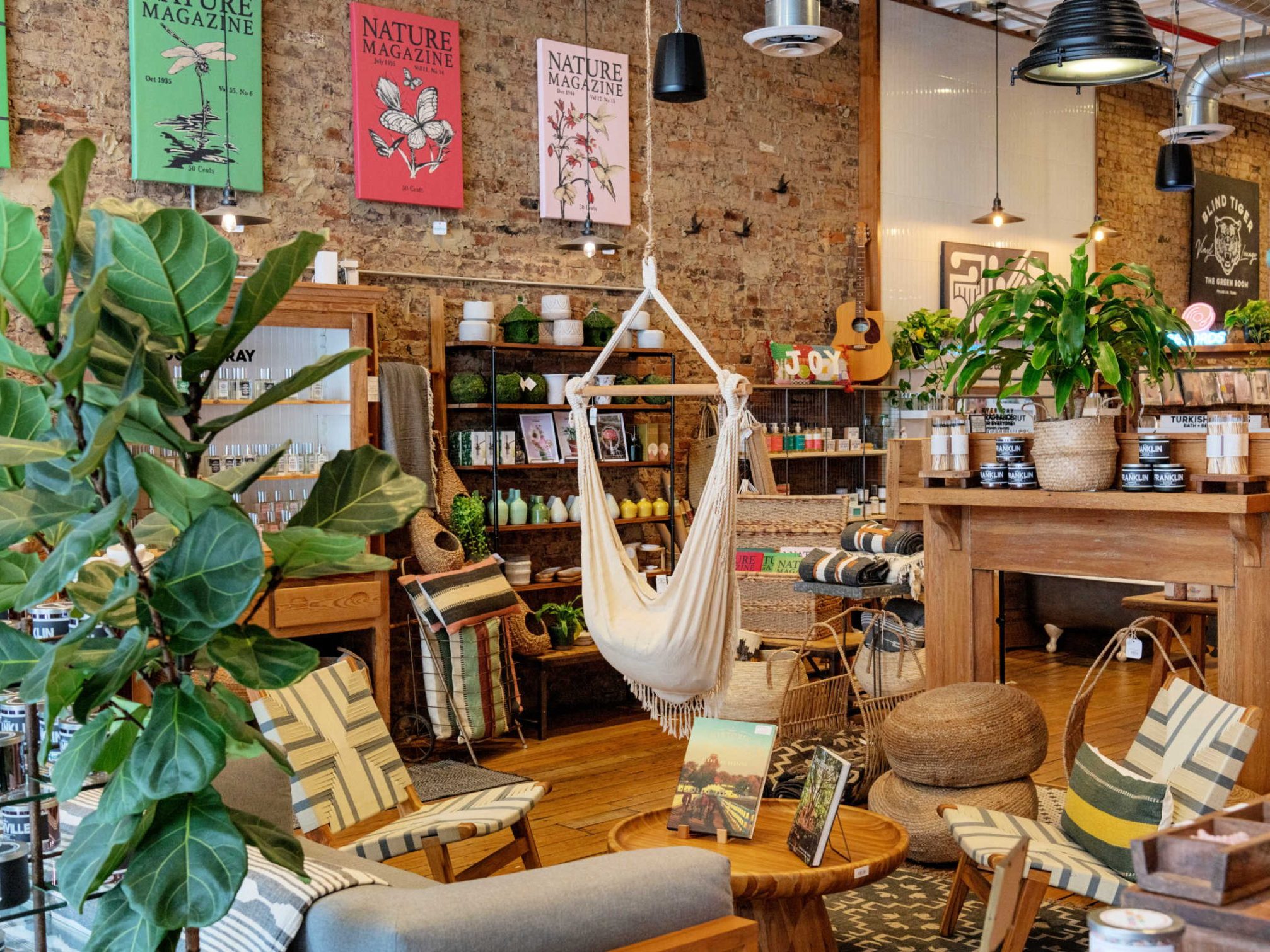 The Green Room Franklin Candles 2048x1511 Interior of a boutique shop displaying plants, woven furniture, and nature-themed wall art on a brick wall