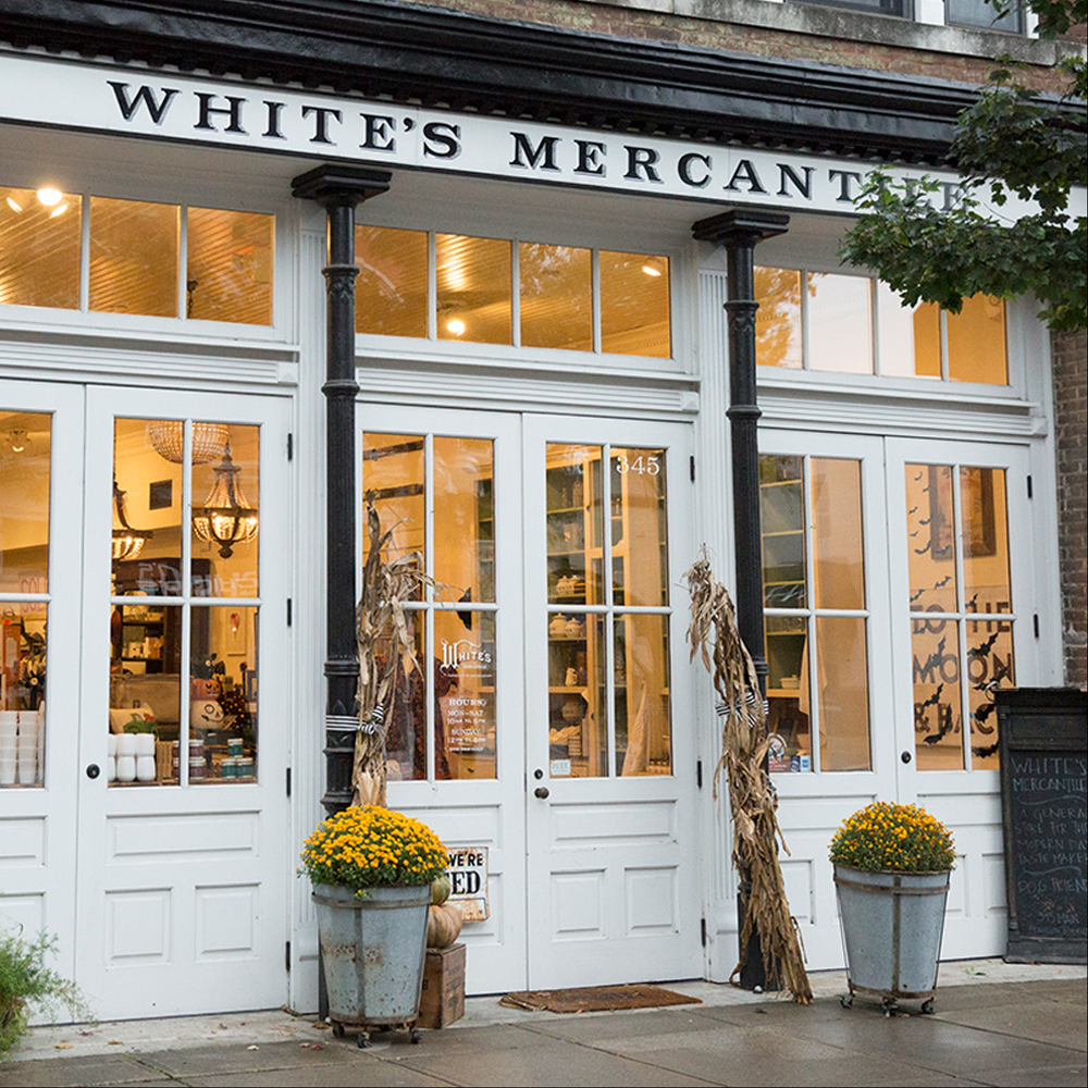 Whites Mercantile Ila Bridal Front of White's Mercantile storefront with glass doors, warm interior lighting, and fall decor (mums in pots, corn stalks, pumpkins).