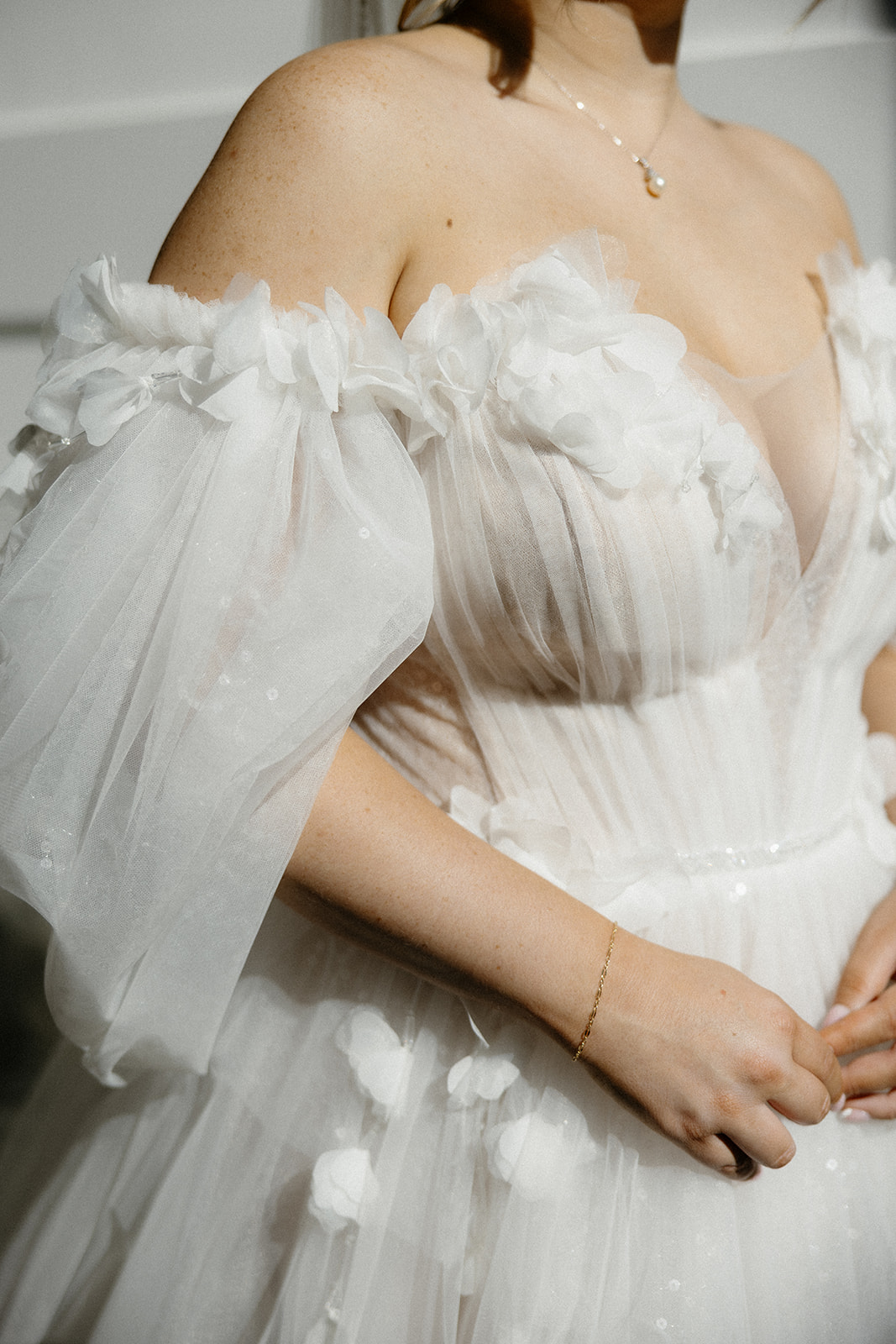 Bride in a white off-the-shoulder wedding gown with sheer puff sleeves and floral appliqués, wearing a necklace and bracelet.