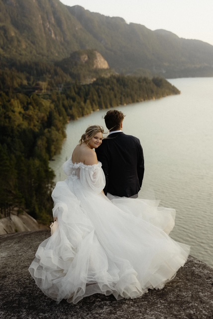 Sylvia Sam Elopement | Ila Bridal Studio Bride in a white off-shoulder gown sits with a groom in a dark suit on a rocky overlook above a lake and mountains.