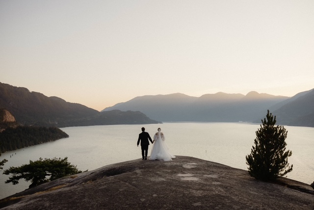 Bride and groom walk hand in hand on a rocky overlook at sunset, with a calm lake and distant mountains in silhouette behind them.